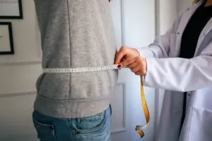 Dietetyk w białym fartuchu mierzy talię ubranej osoby taśmą krawiecką