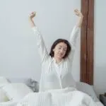 Kobieta przeciąga się w łóżku po przebudzeniu, na szafce nocnej stoi budzik