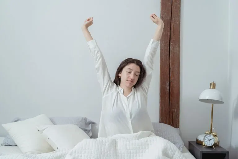 Kobieta przeciąga się w łóżku po przebudzeniu, na szafce nocnej stoi budzik